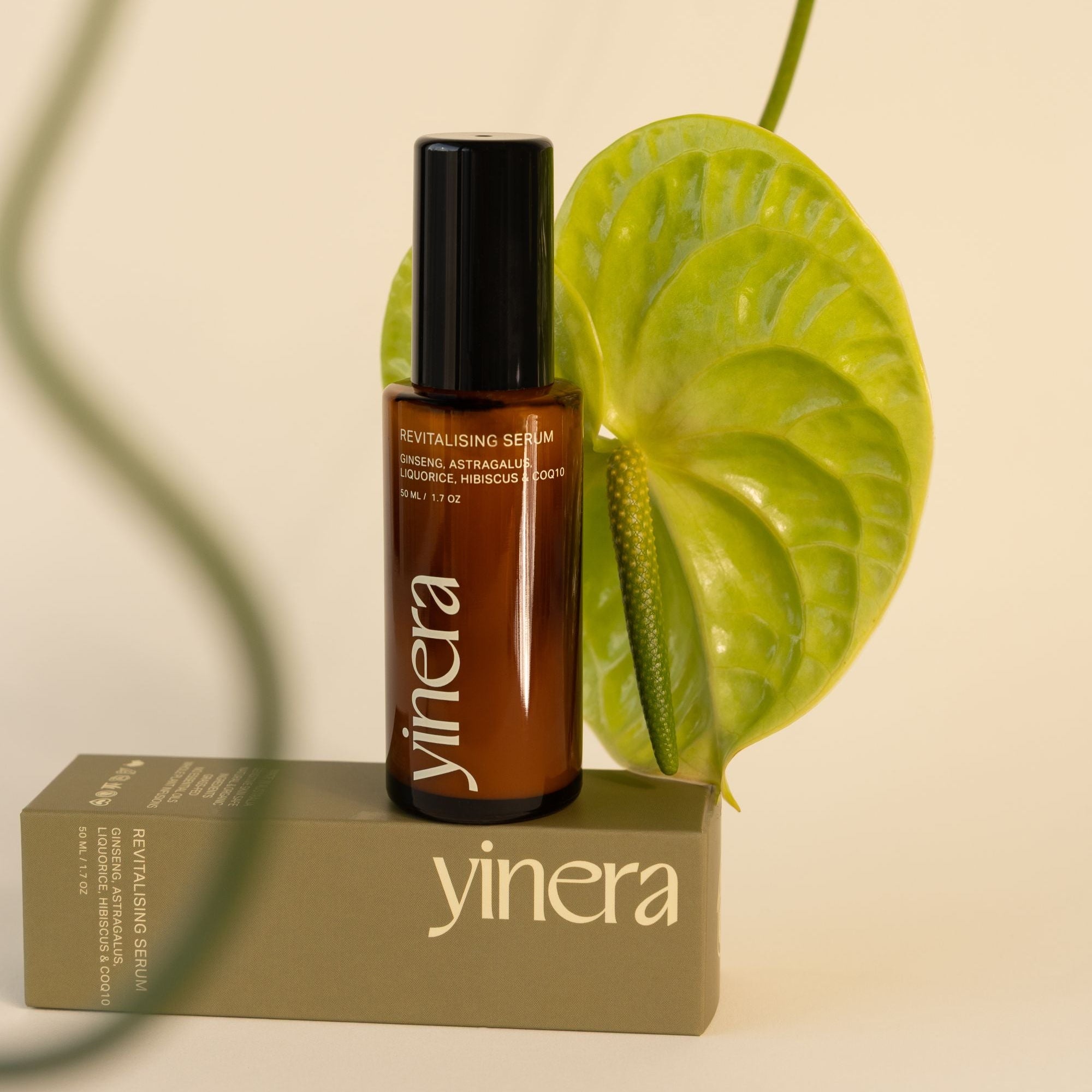 Brown serum bottle with black cap on a beige box, surrounded by green leaves on a beige background yinera skincare